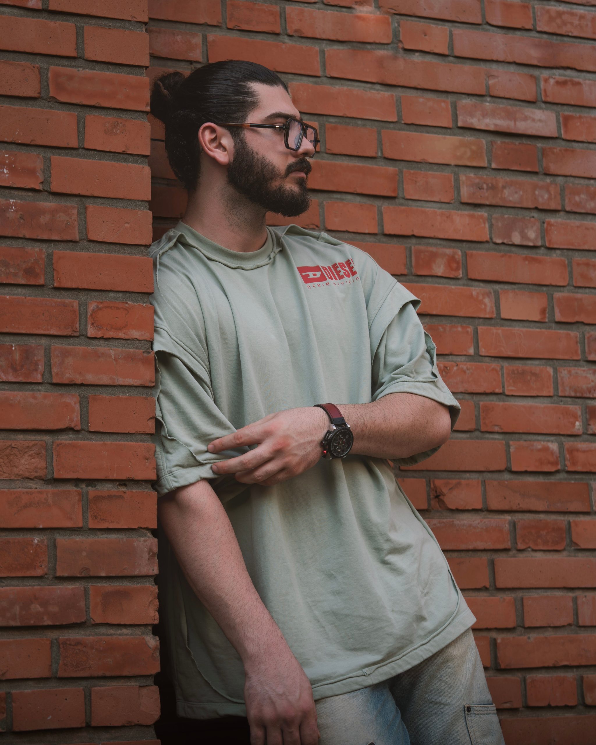 Fashionable man with sunglasses and beard leans casually against a brick wall in an urban setting.