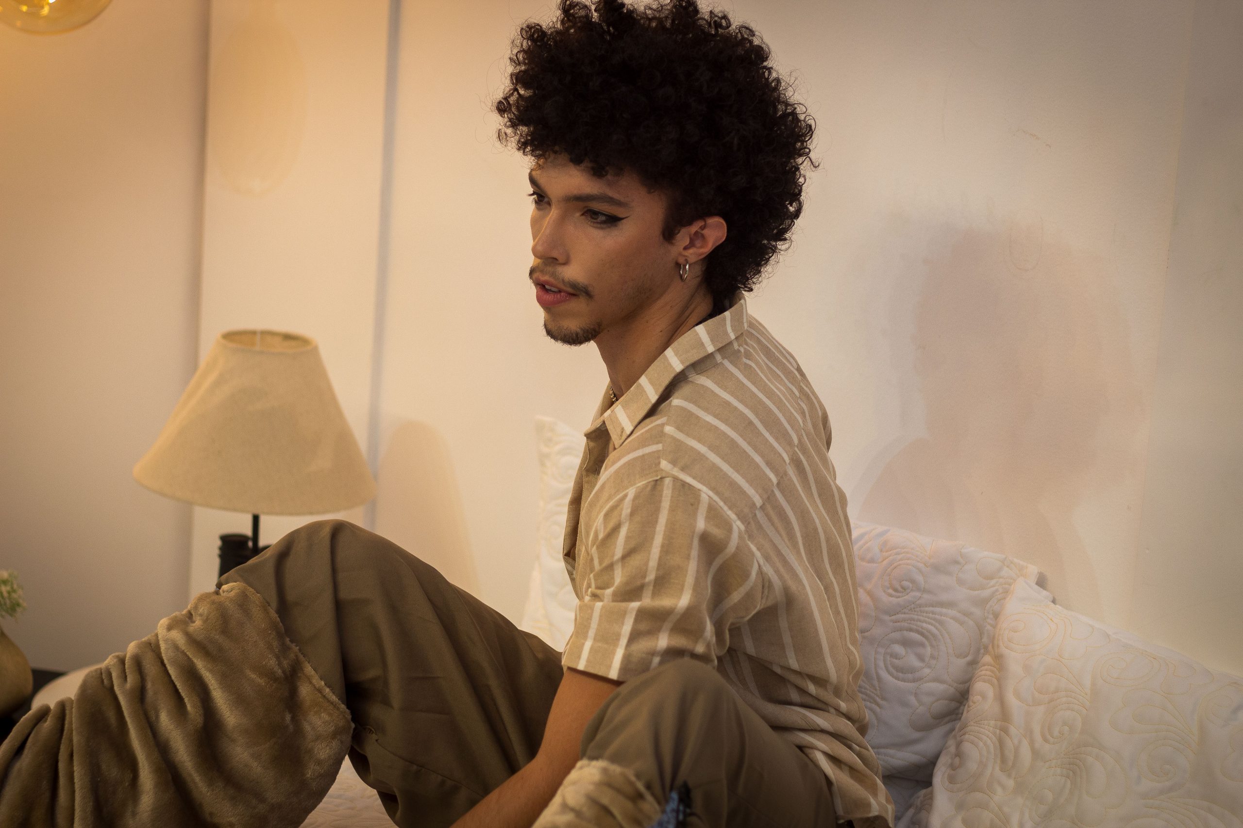 A young man sits thoughtfully on a bed in a warm, cozy bedroom with soft lighting.